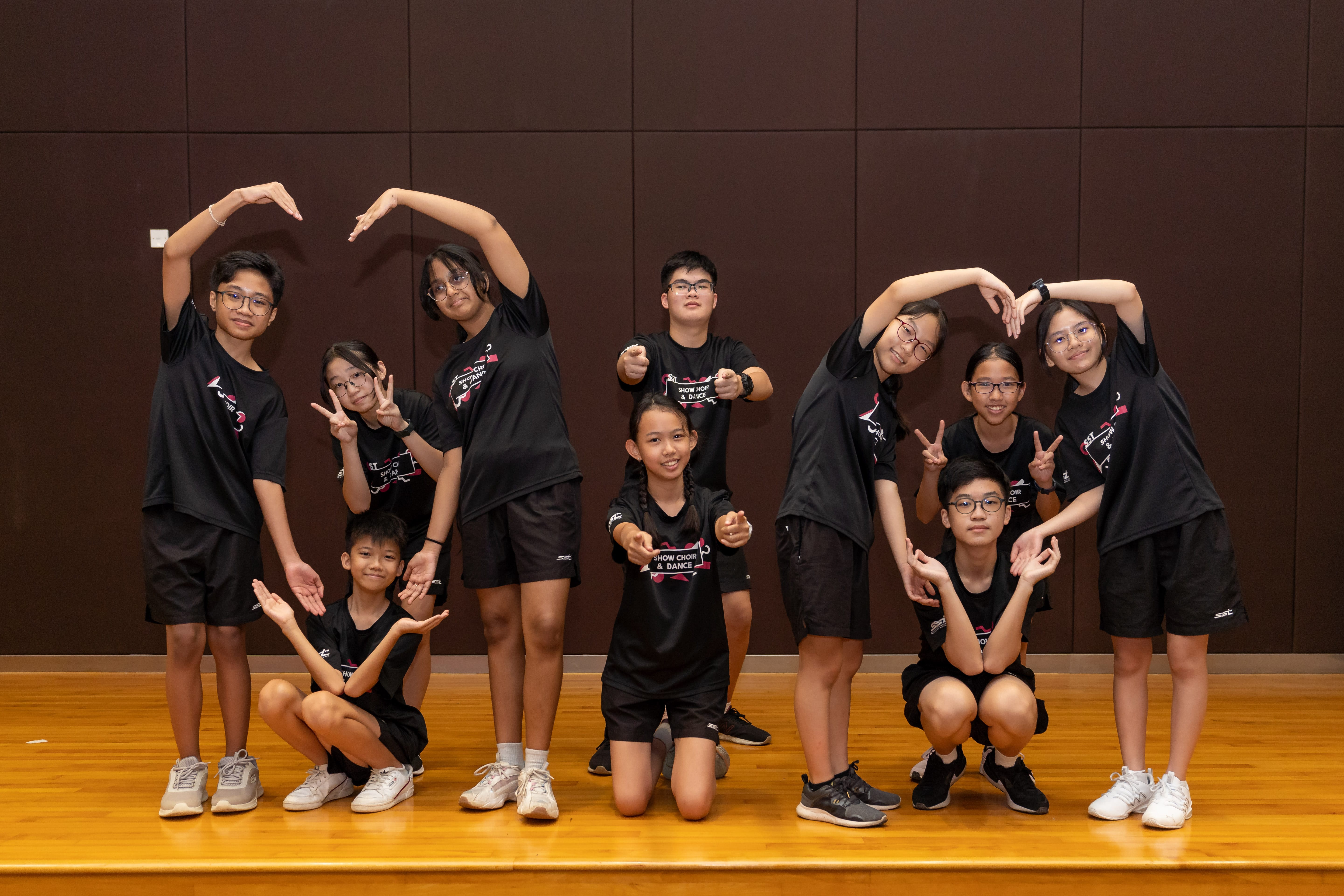 Eleven children in matching black Show Choir and Dance uniforms pose with fun gestures on a wooden stage.